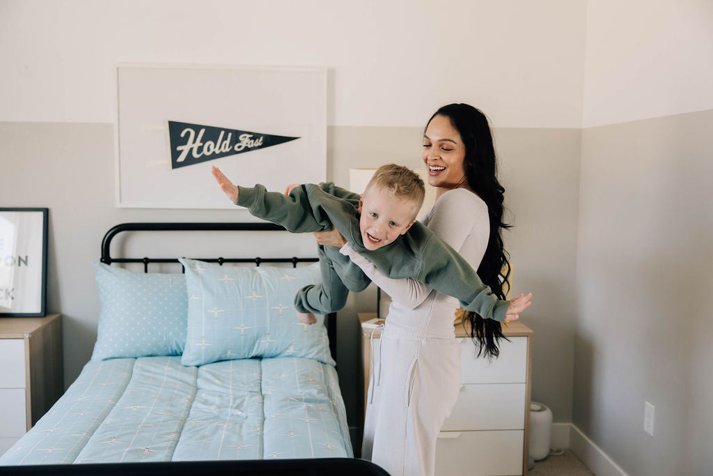 A mom holding her son with his arms out, pretending he's an airplane, standing beside a twin bed made with blue airplane themed zipper bedding.