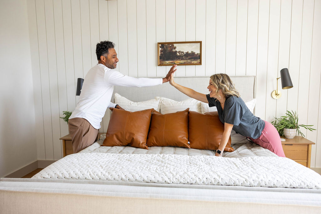 A couple leaning over a freshly made bed, giving each other a high five to celebrate having made the bed together.