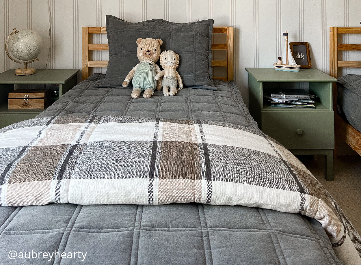 A child's twin bed made with gray grid pattern zipper bedding, a plaid blanket folded at the foot of the bed and 2 stuffed animals propped against the pillow.