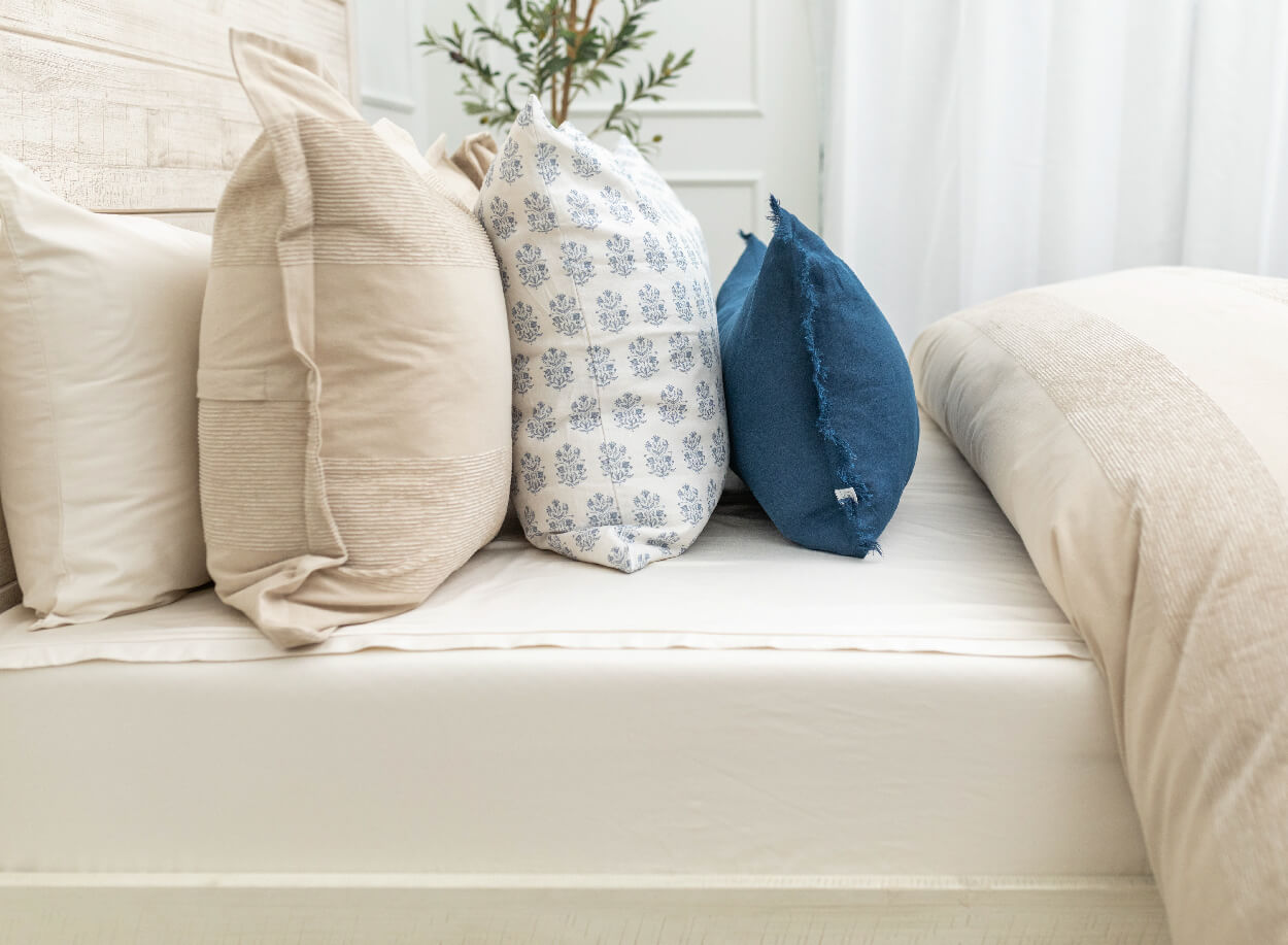 A side shot of a queen med made with ivory zipper sheets and a tan colored duvet, an assortment of tan and blue throw pillows arranged on top.