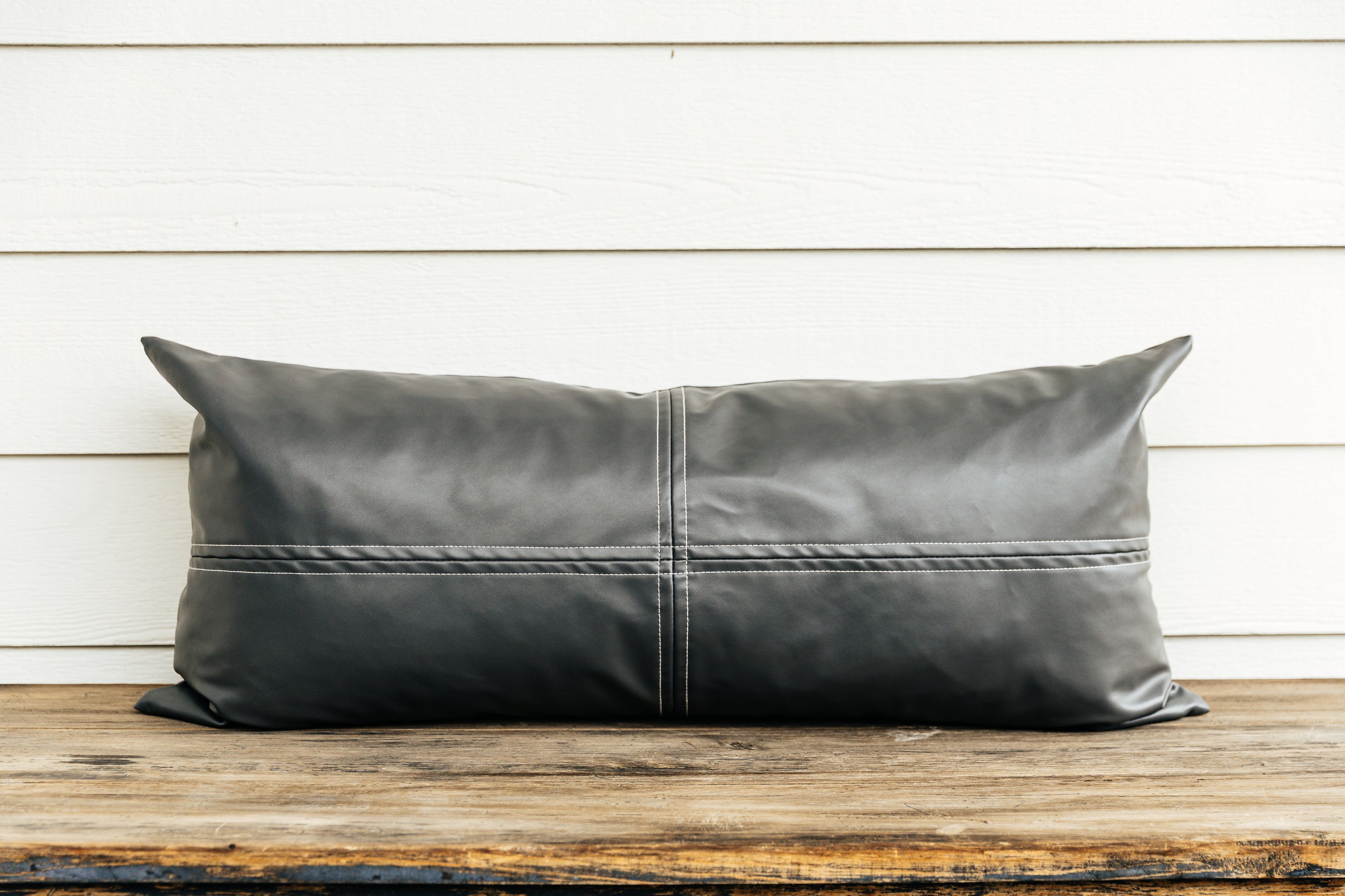 A faux leather charcoal lumbar pillow sitting on a brown bench with a white backdrop.