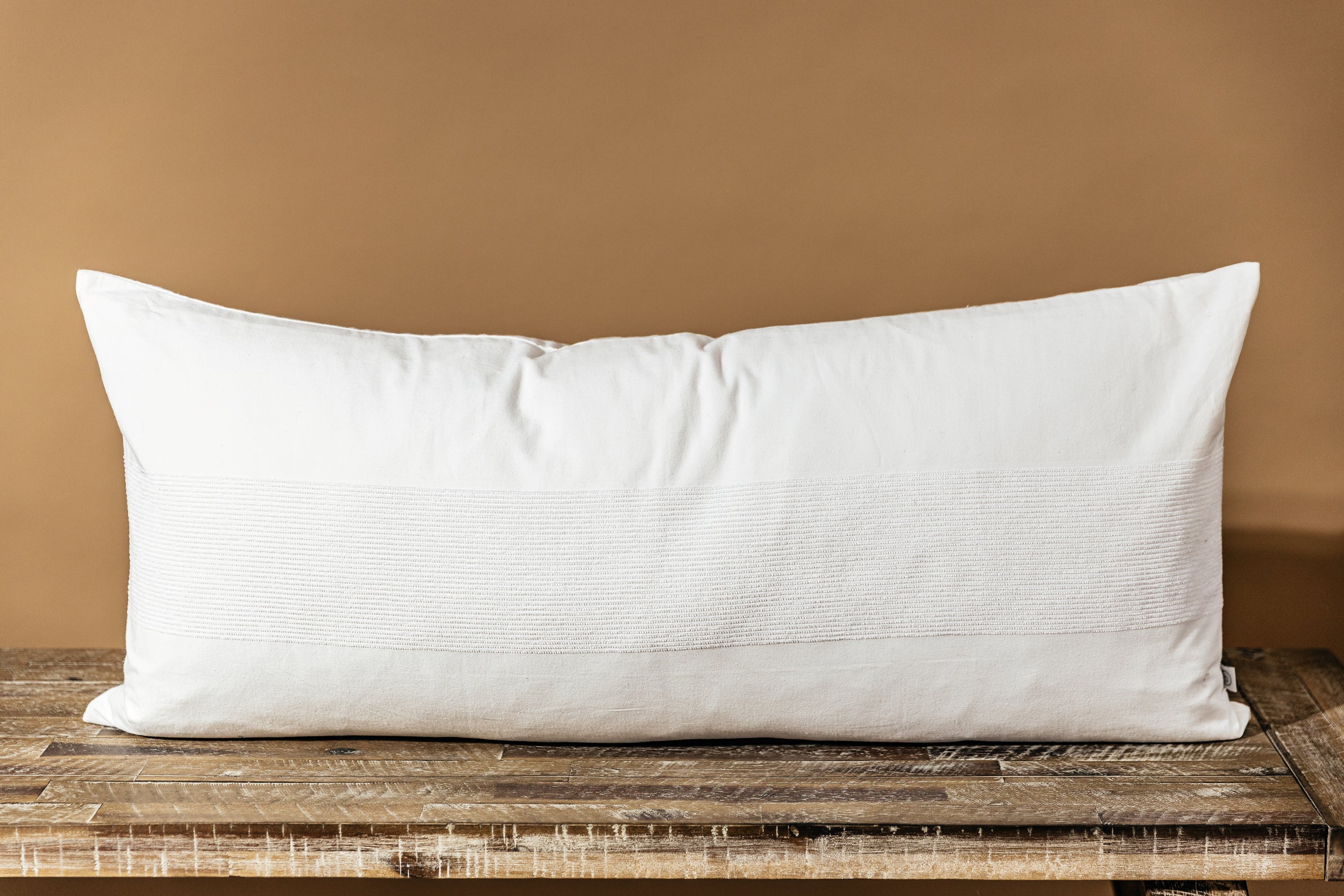 A white lumbar pillow on a wood bench in front of a brown backdrop.