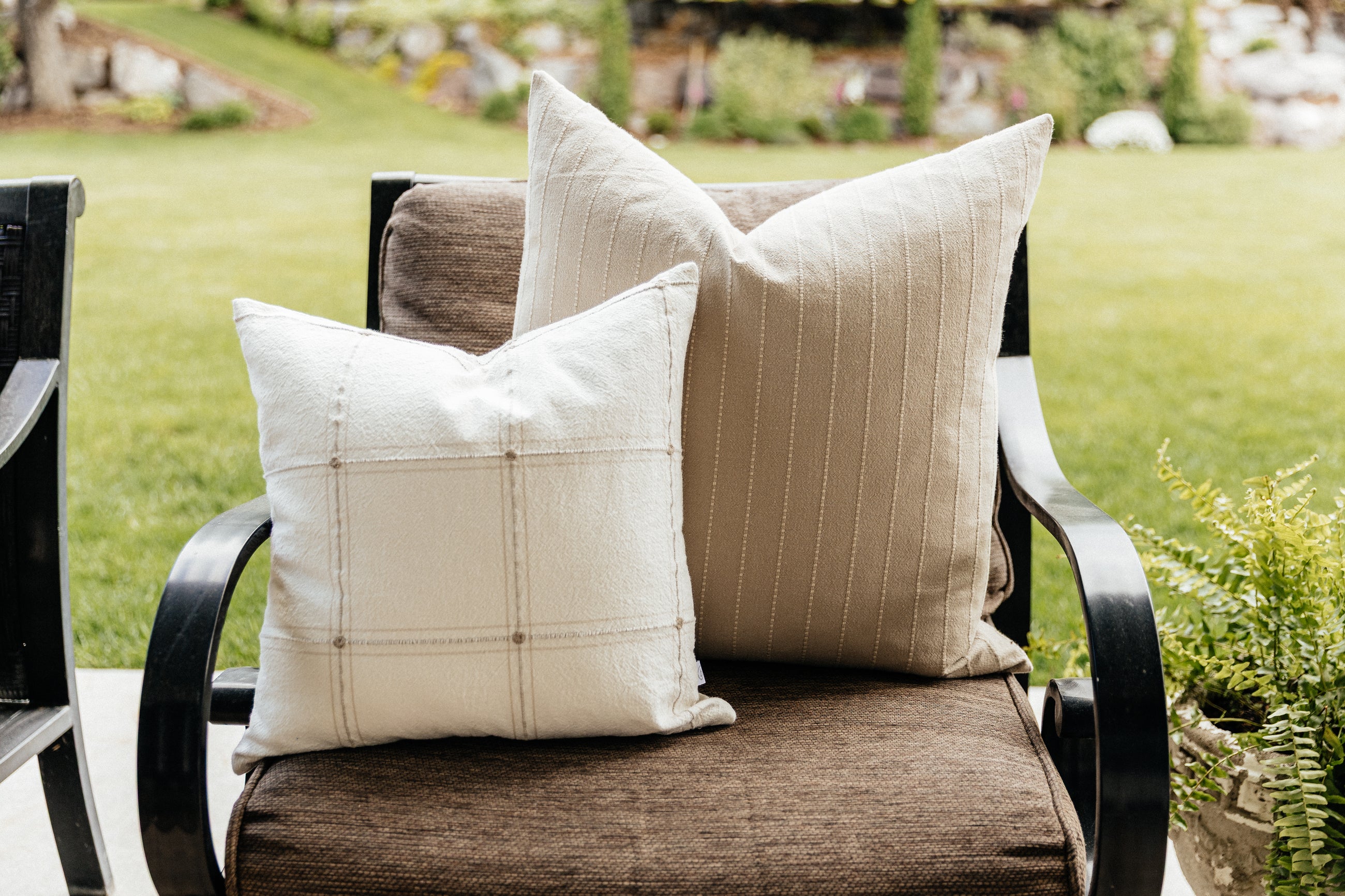 Two neutral pillows arranged on an outdoor chair on a back patio, a lush green lawn in the background.