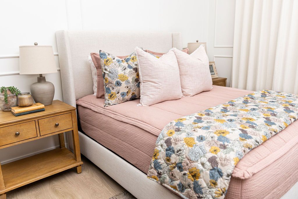 A brightly lit bedroom with a bed made with woven pink zipper bedding and an assortment of pink and floral tan pattern pillows and blanket.