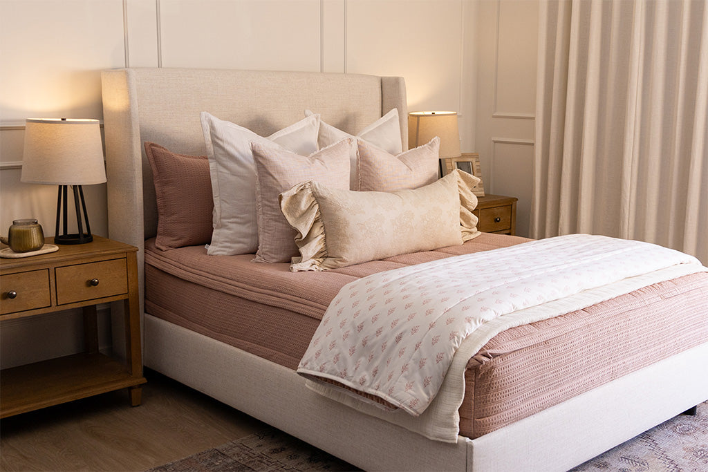 A queen bed made with woven pink zipper bedding and an assortment of neutral and light pink throw pillows and floral blanket.