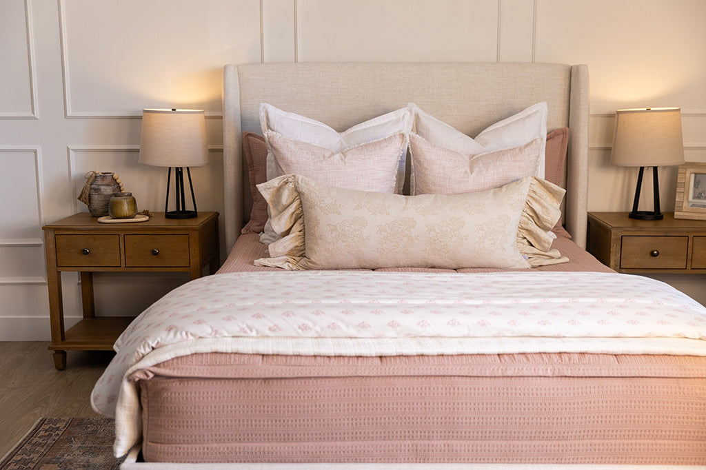 A queen bed made with woven pink zipper bedding and an assortment of neutral and light pink throw pillows and floral blanket.