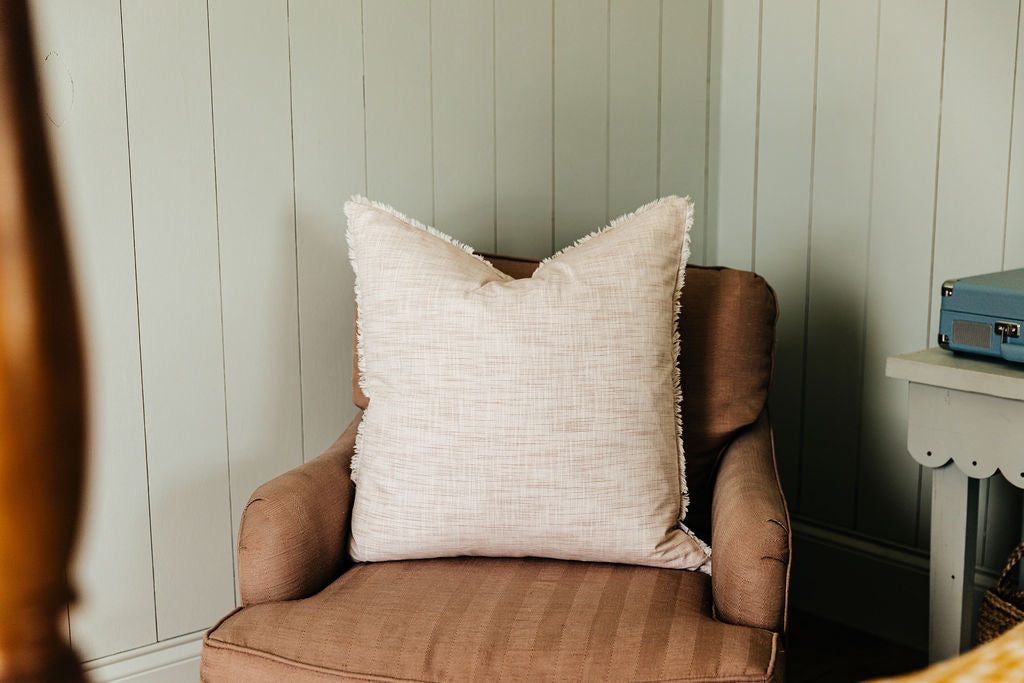A light pink woven striped euro pillow arranged on a brown chair in a corner of a bedroom.