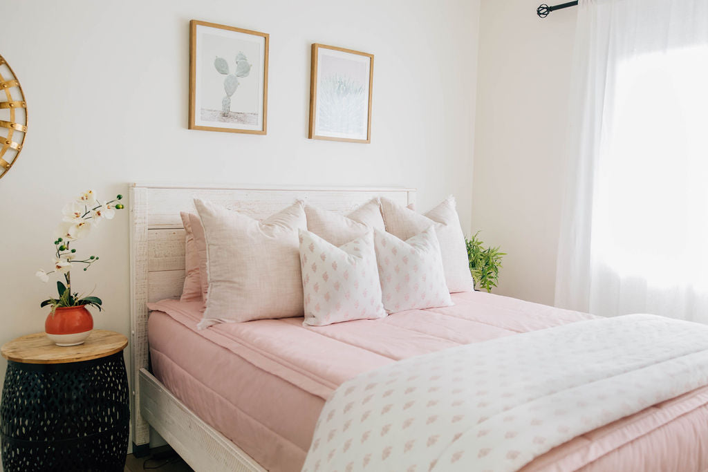 A brightly lit bedroom with a bed made with pink zipper bedding and light pink and floral pillows and blanket.
