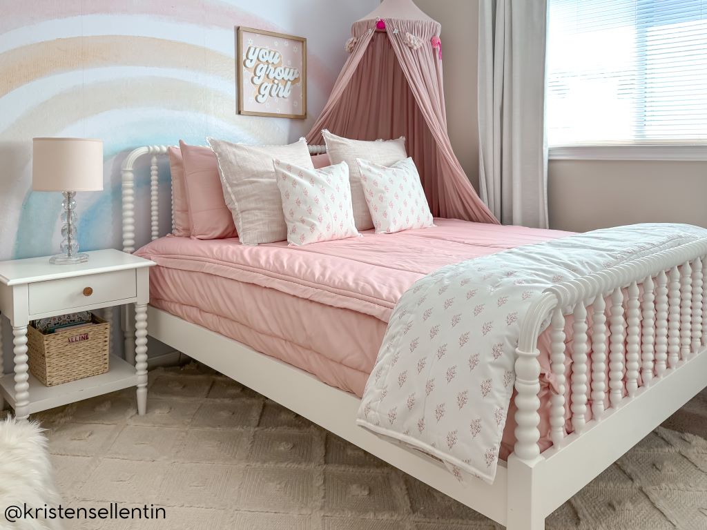 A bed made with pink zipper bedding and an assortment of light pink and floral pillows and blanket with pink canopy hanging over the side of the bed.