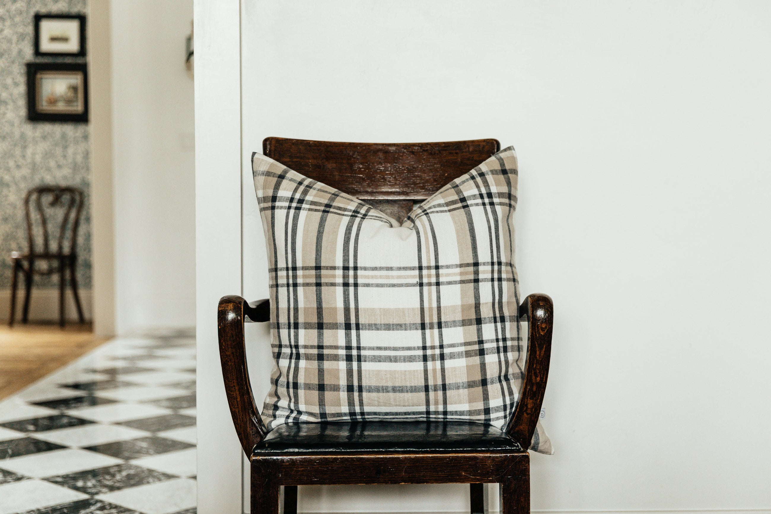 A charcoal, cream, and tan plaid euro pillow on a brown wood chair against a white wall.
