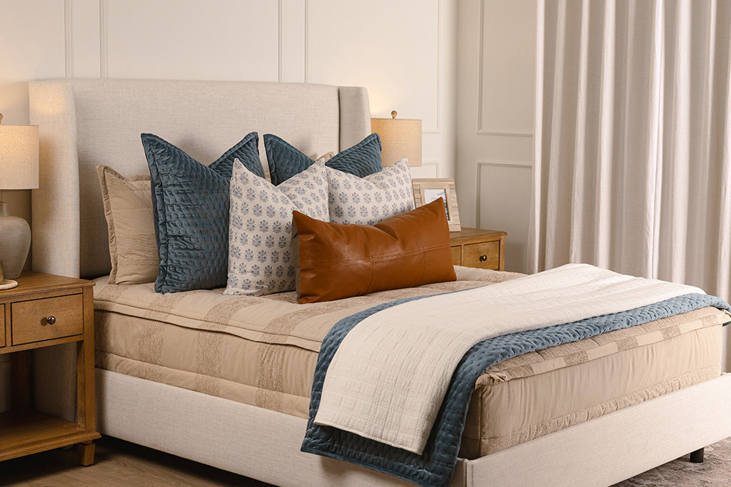 A queen bed made with tan zipper bedding and an assortment of blue, leather, and white bedding accessories.