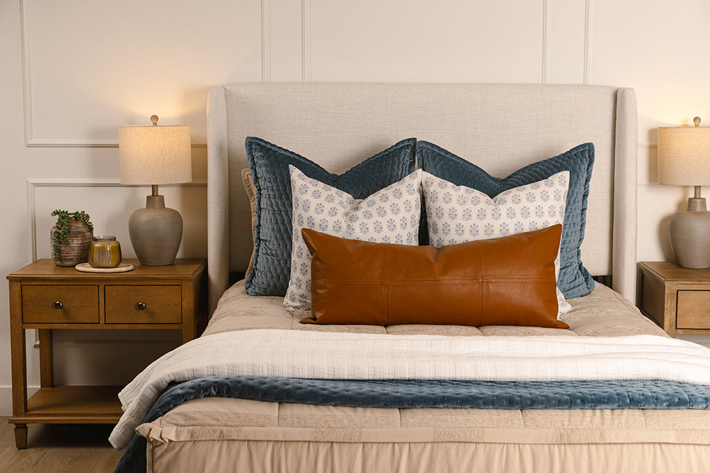 A queen bed made with tan zipper bedding and an assortment of blue, leather, and white bedding accessories.