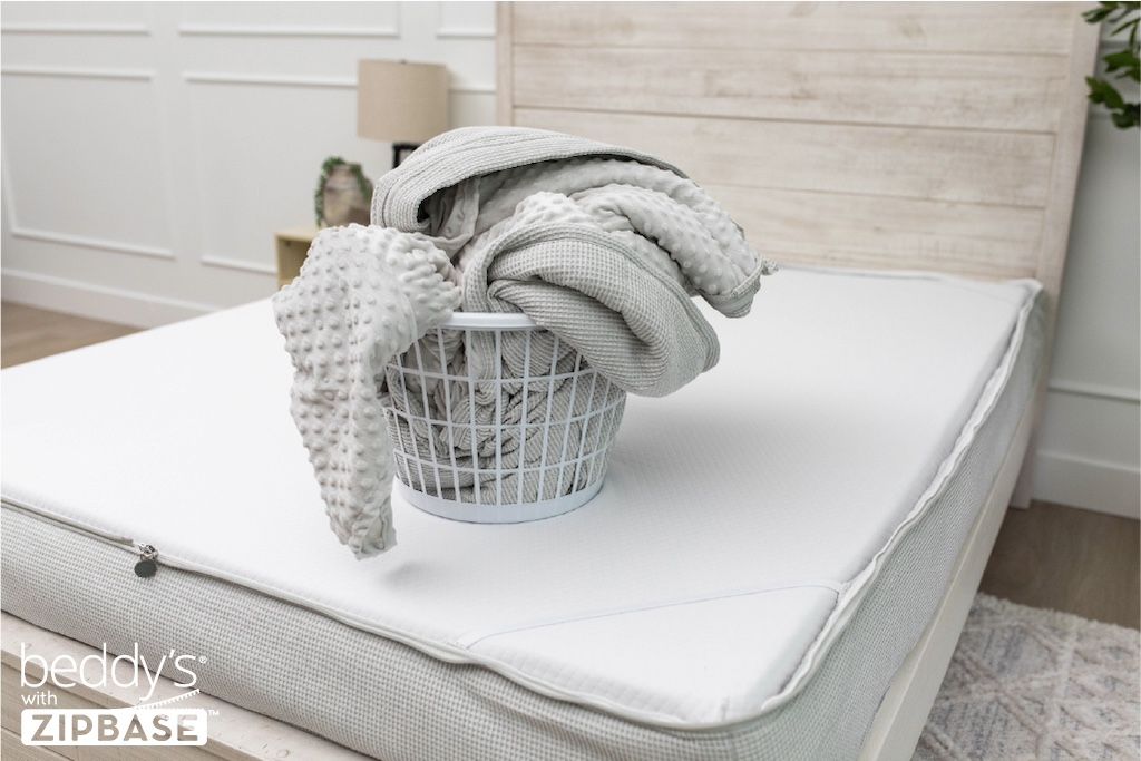 A bed made with just the fitted bedding base around the sides of the mattress, with the top comforter and ZipBase sheet in a basket on top of the bed.