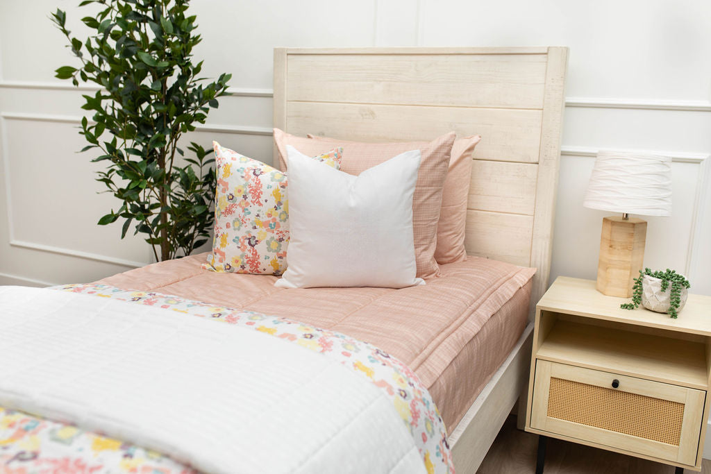 A twin bed made with peach pink zipper bedding and adorned with neutral and multicolor floral pattern pillows and blankets.