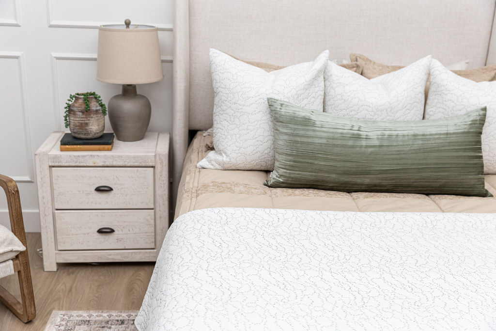 A bed made with tan zipper bedding with white stitched throw pillows, a green lumbar pillow, and a white stitched throw blanket.