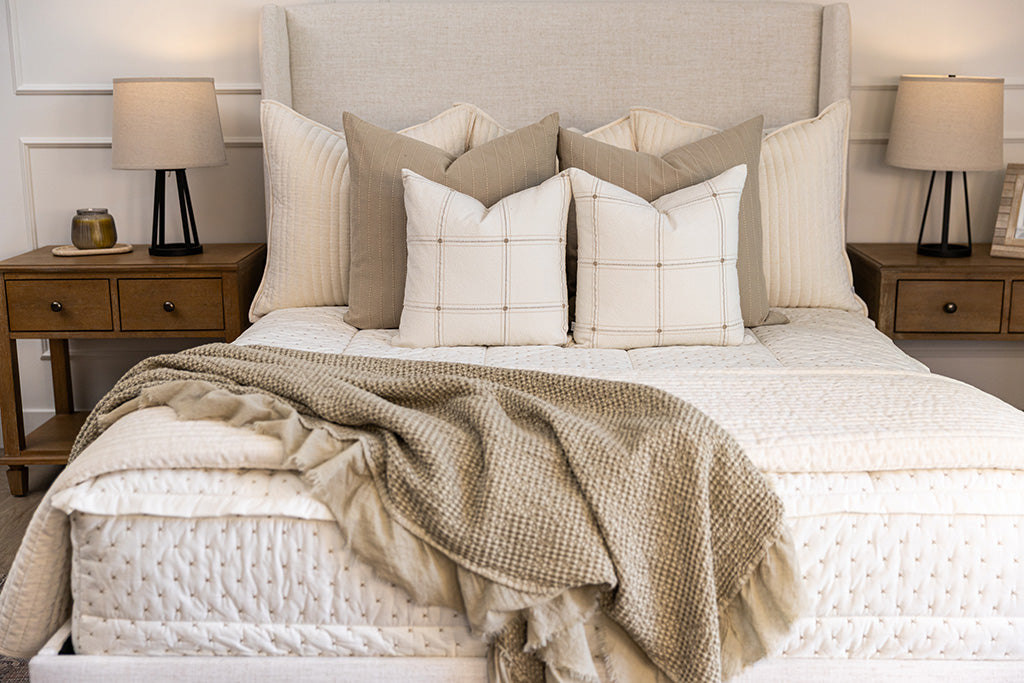 A lightly lit bedroom with a bed made with cream embroidered zipper bedding, decorated with neutral throw pillows and blankets.