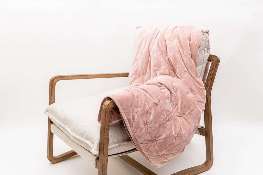 A cushioned chair with a wood frame, a velvet pink blanket draped across the arm and back of the chair.