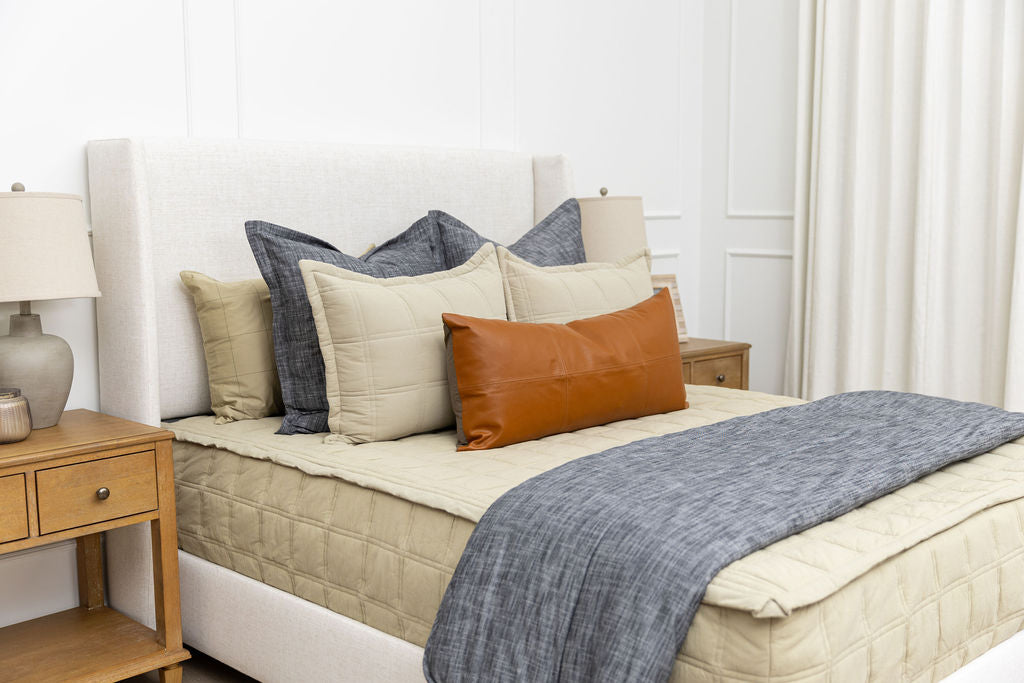 A bed made with khaki green zipper bedding and an assortment of neutral and dark gray pillows and blanket.