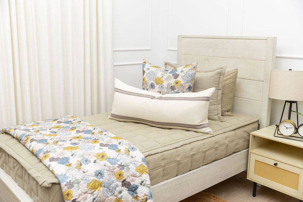 A twin bed made with khaki green zipper bedding and an assortment of floral and cream striped pillows and blanket.