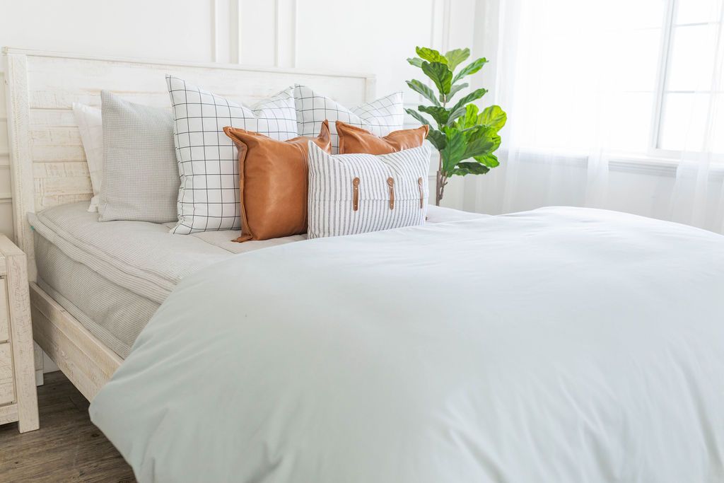 A brightly lit bedroom with a bed made with light gray waffle textured zipper bedding and a light blue zipper duvet folded half way down the bed and adorned with neutral and leather pillows