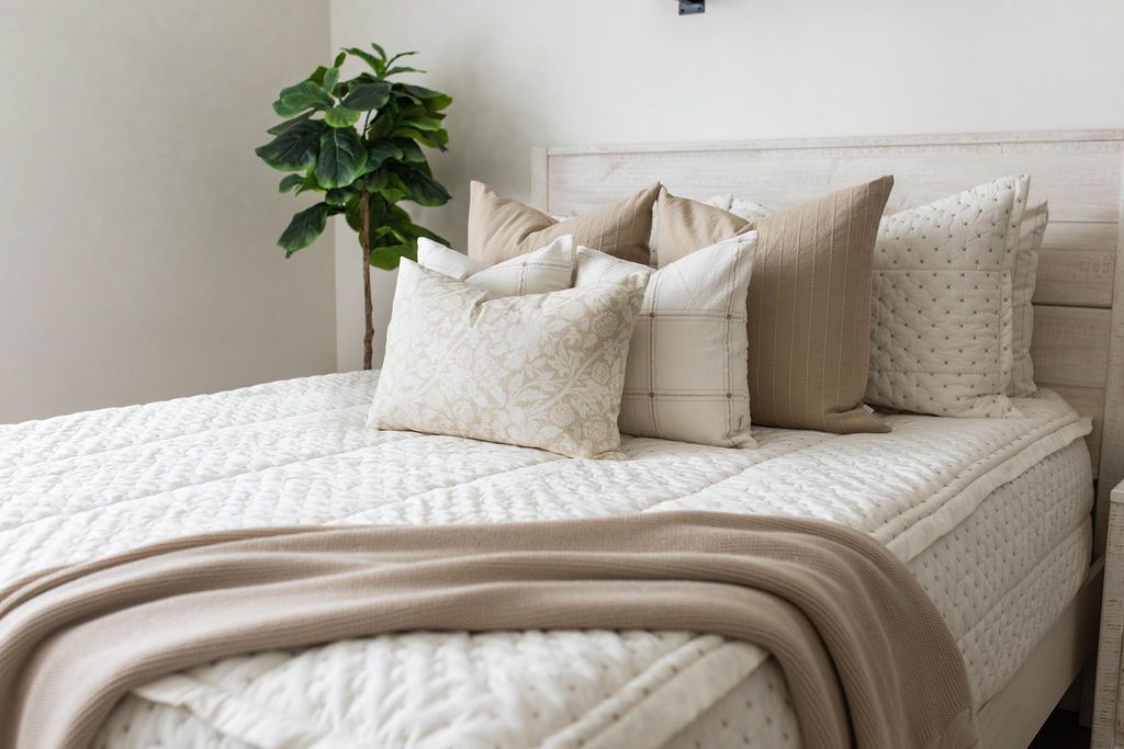 A queen bed made with cream embroidered zipper bedding and neutral pattern tan pillows and matching woven throw blanket at the end of the bed.