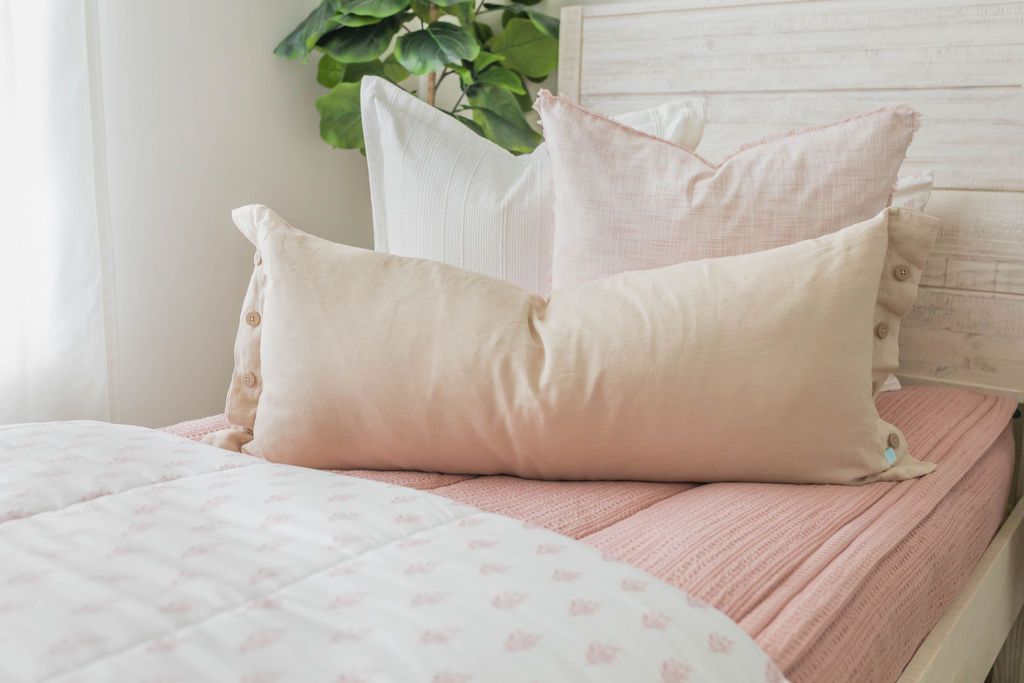 A close up view of a twin bed made with pink woven zipper bedding and neutral and light pink pillows and a floral pattern blanket.