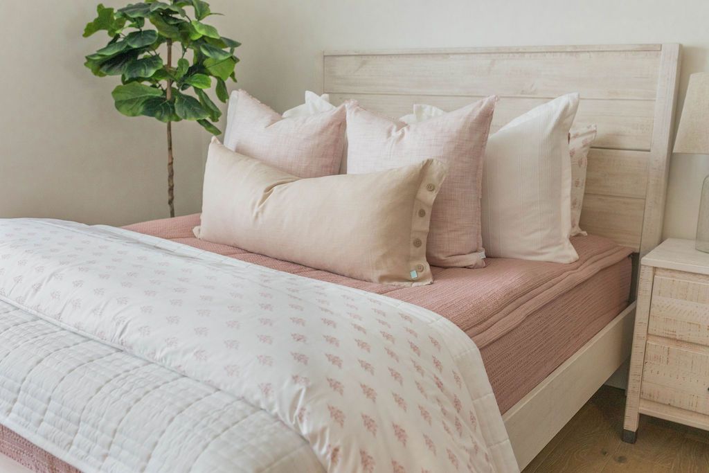 A queen bed made with pink woven zipper bedding and neutral and pink pillows and coordinating blankets at the end of the bed.