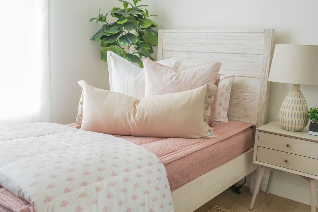 A twin bed made with pink woven zipper bedding and neutral and pink pillows and a floral design white blanket.