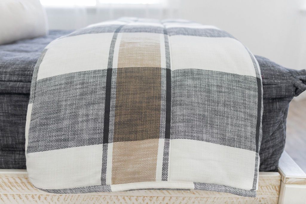 A close up side view of a bed made with deep gray zipper bedding and a tan, cream, and gray plaid pattern blanket thrown over the side of the bed.