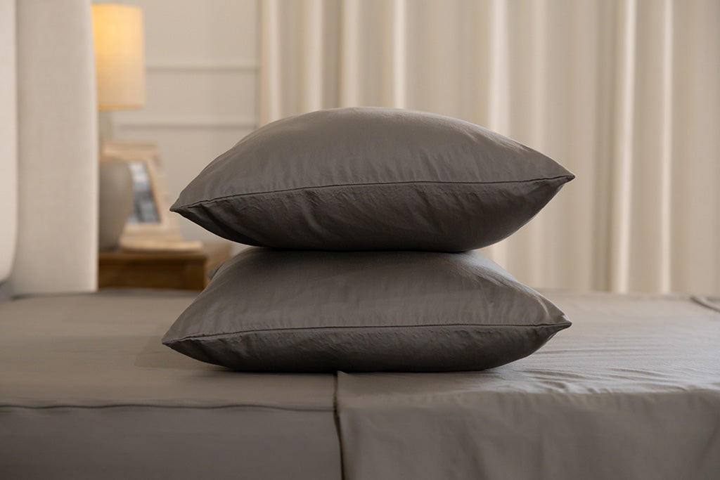 A side view of a bed made with gray zipper sheets and the top sheet slightly unzipped and folded with the matching pillows stacked on top of each other.