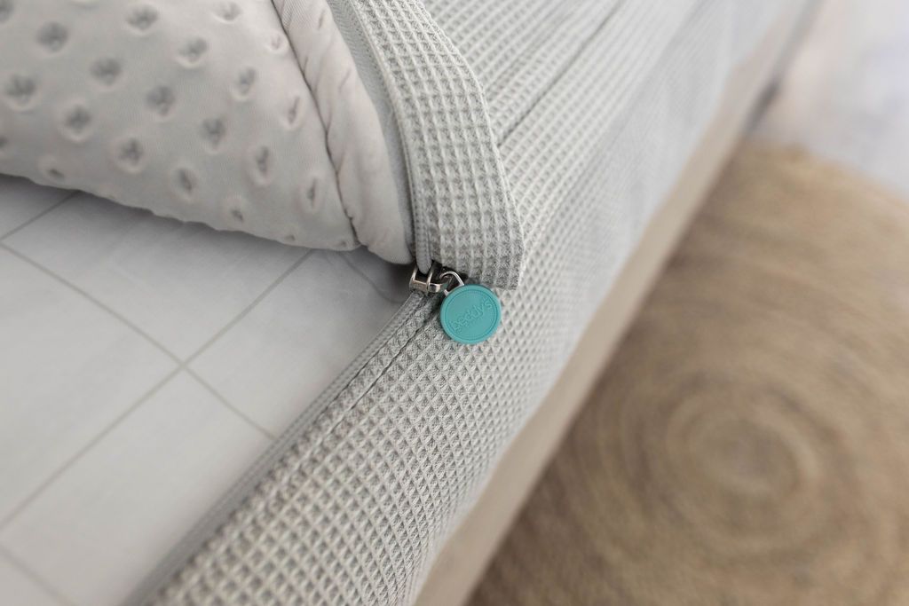 A close up view of a bed made with light gray waffle textured zipper bedding with the bed slightly unzipped to reveal the minky comforter and the white and gray grid pattern bottom cotton lining.