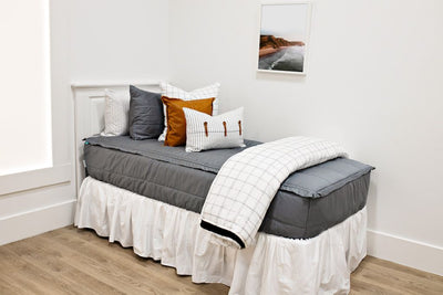 White twin size bed with gray bedding, a white and black grid euro pillow, a medium brown leather pillow, a white and black striped lumbar pillow, a white bed skirt and a white and black grid blanket at the foot of the bed.