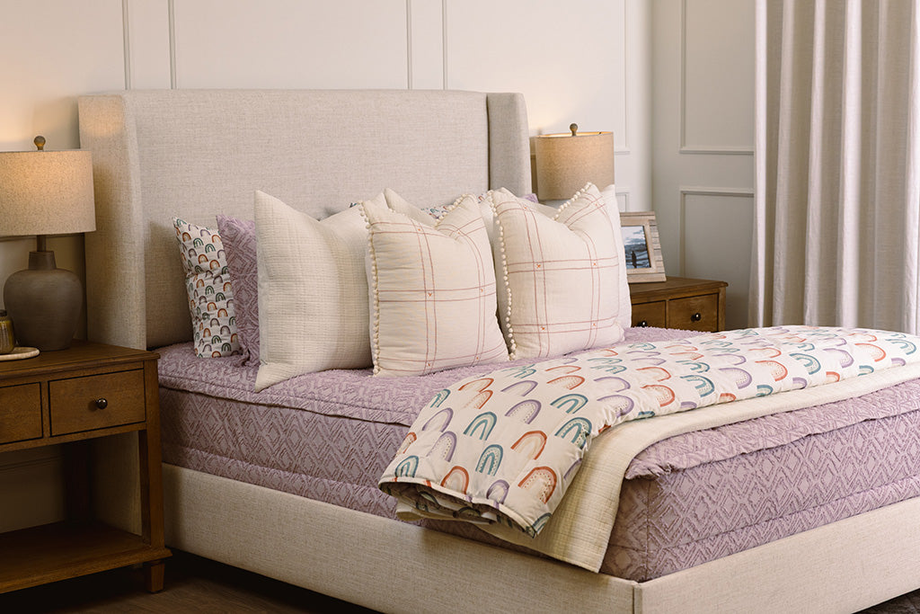 White queen bedframe purple textured zipper bedding and textured euro pillow with tassels, pastel rainbow pillow, and a white blanket with ombre purple, orange and green rainbows