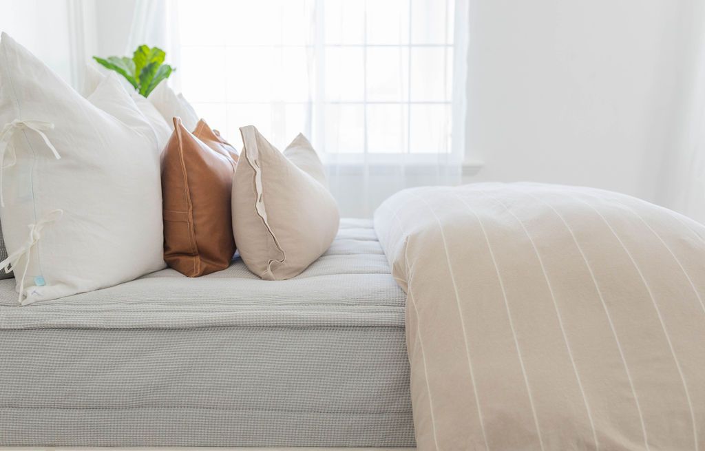 A side view of a bed made with light gray waffle textured zipper bedding and a tan and white striped zipper duvet folded half way down the bed and adorned with neutral pillows.