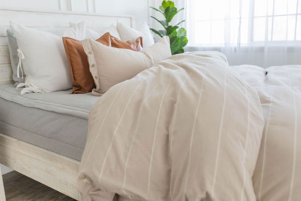 A brightly lit bedroom with a bed made with light gray waffle textured zipper bedding and a tan and white striped zipper duvet partially folded half way down the bed and adorned with neutral pillows.