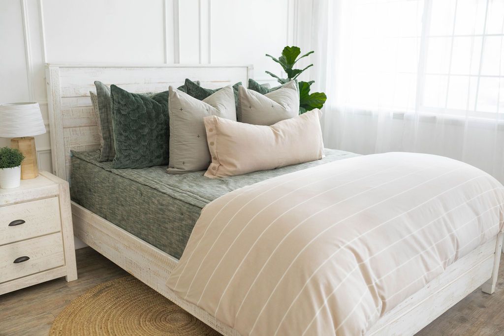 A brightly lit bedroom with a bed made with deep emerald green zipper bedding and a tan and white striped zipper duvet folded half way down the bed and adorned with neutral and green pillows.