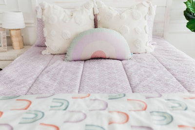 purple textured bedding with textured euro pillow with tassels, pastel rainbow pillow, and a white blanket with ombre purple, orange and green rainbows