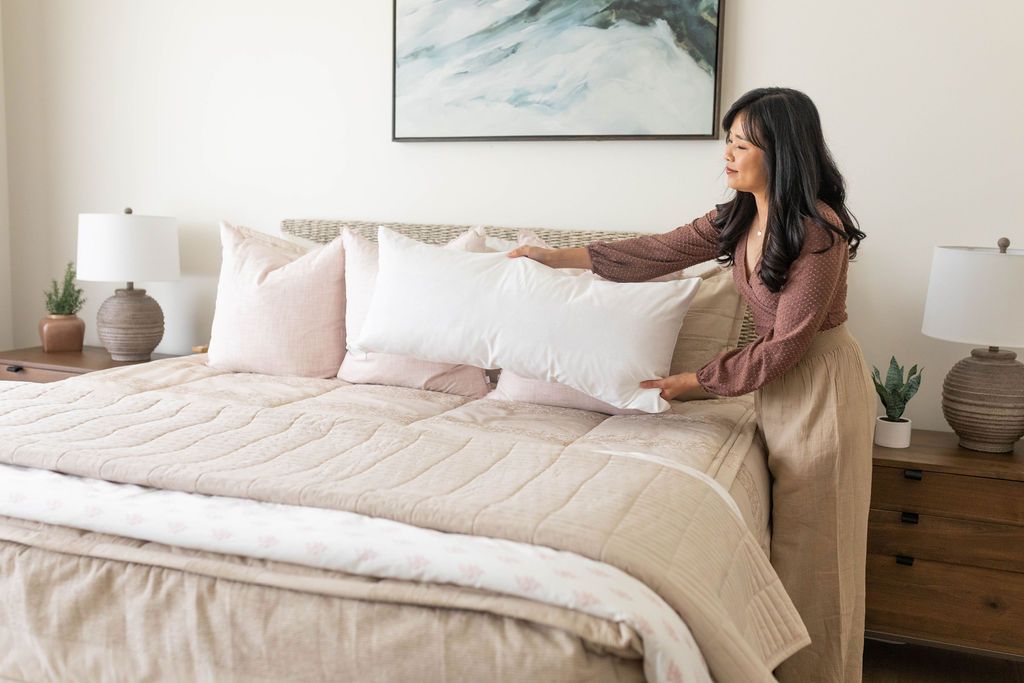 A bed made with tan faded stripes zipper bedding and an assortment of neutral and light pink pillows and blankets with a woman placing a large white lumbar pillow onto the bed.