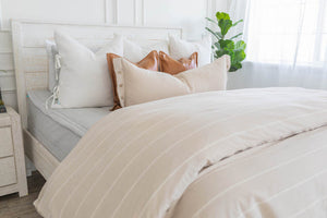 A bed made with light zipper bedding with a duvet folded at the foot of the bed and an assortment of throw pillows.