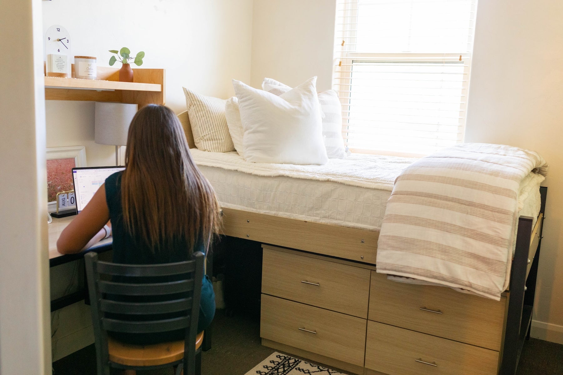 
      A college student sitting at her desk in her dorm room next to her bed that has zipper bedding, a striped blanket, and white throw pillows on it.
    