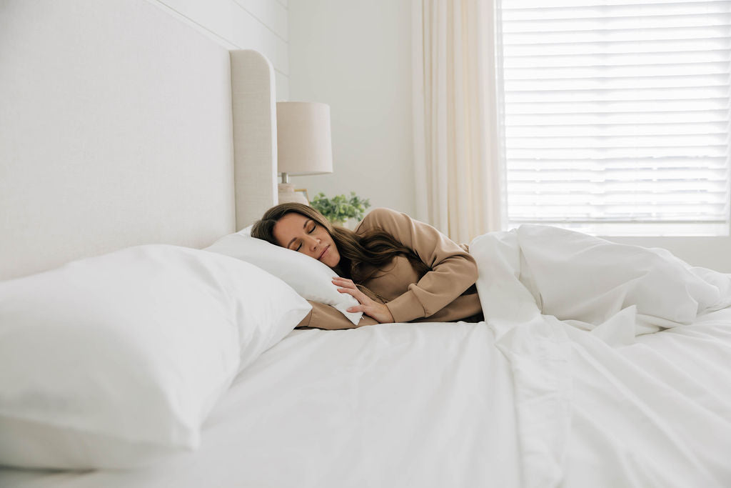 
      A woman asleep in a bed made with crisp white organic bedding, with natural light coming through the window,
    