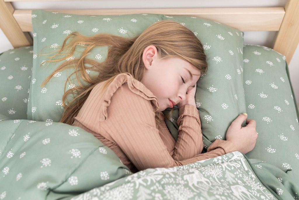 
      A young girl with long blonde hair asleep in a bed made with sage green woodland animal zipper bedding.
    