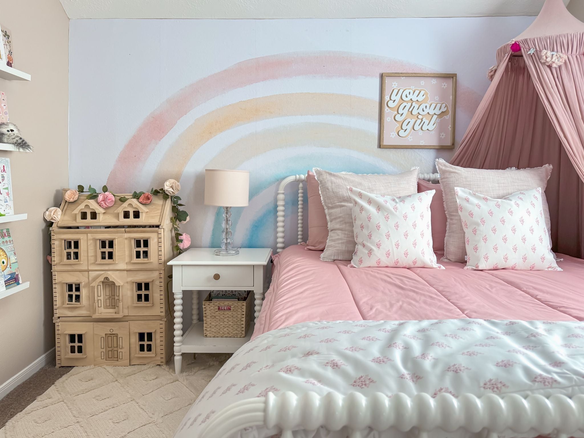 
      A cozy, pastel-themed girl's bedroom featuring pink Beddy's bedding, patterned pillows, a nightstand, a wooden dollhouse adorned with pink flowers, and a whimsical rainbow mural on the wall.
    