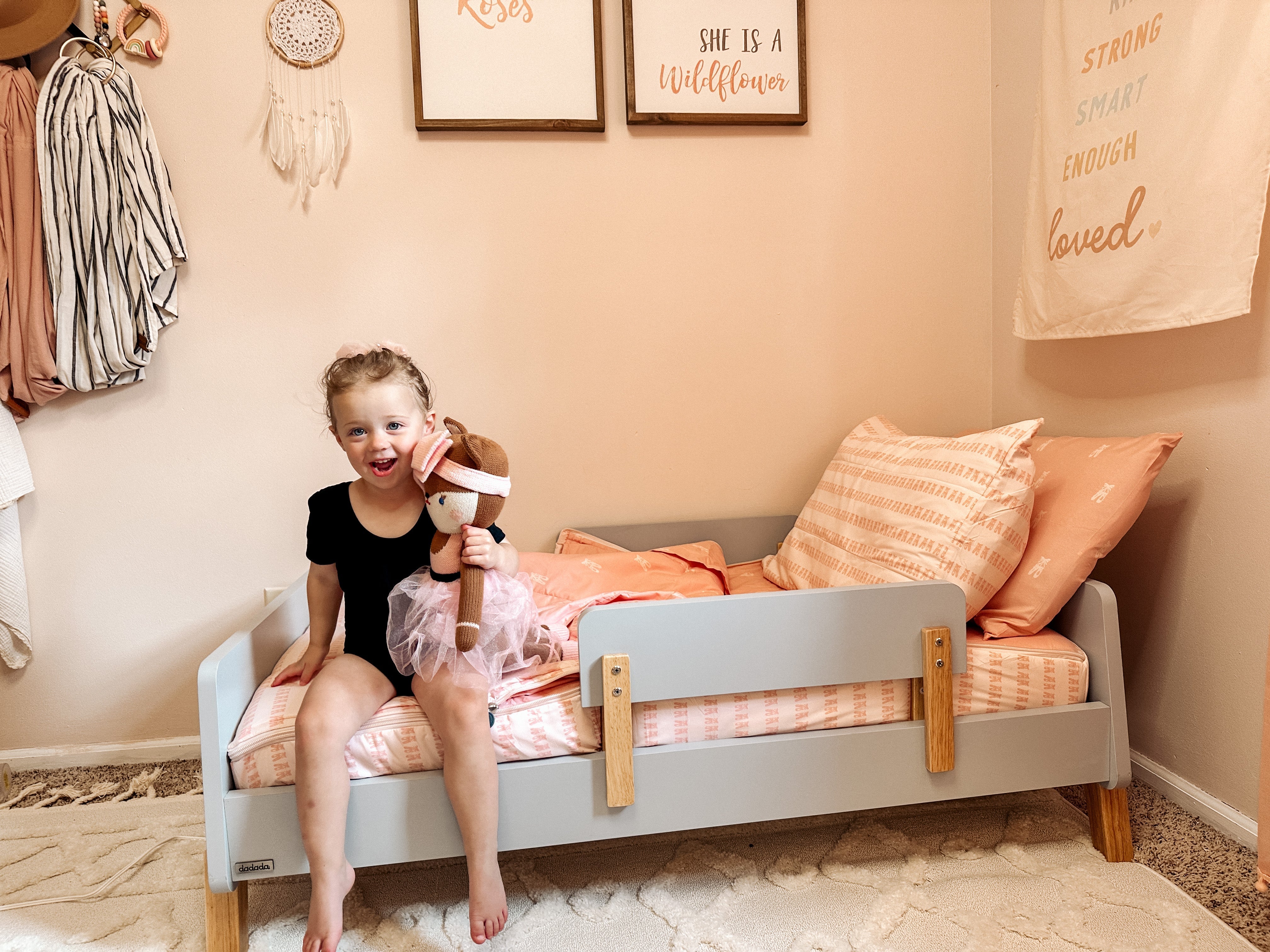 
      A young girl in a black leotard and holding a stuffed ballerina doll, sitting on her toddler sized bed, which is made with ballet slipper printed zipper bedding.
    