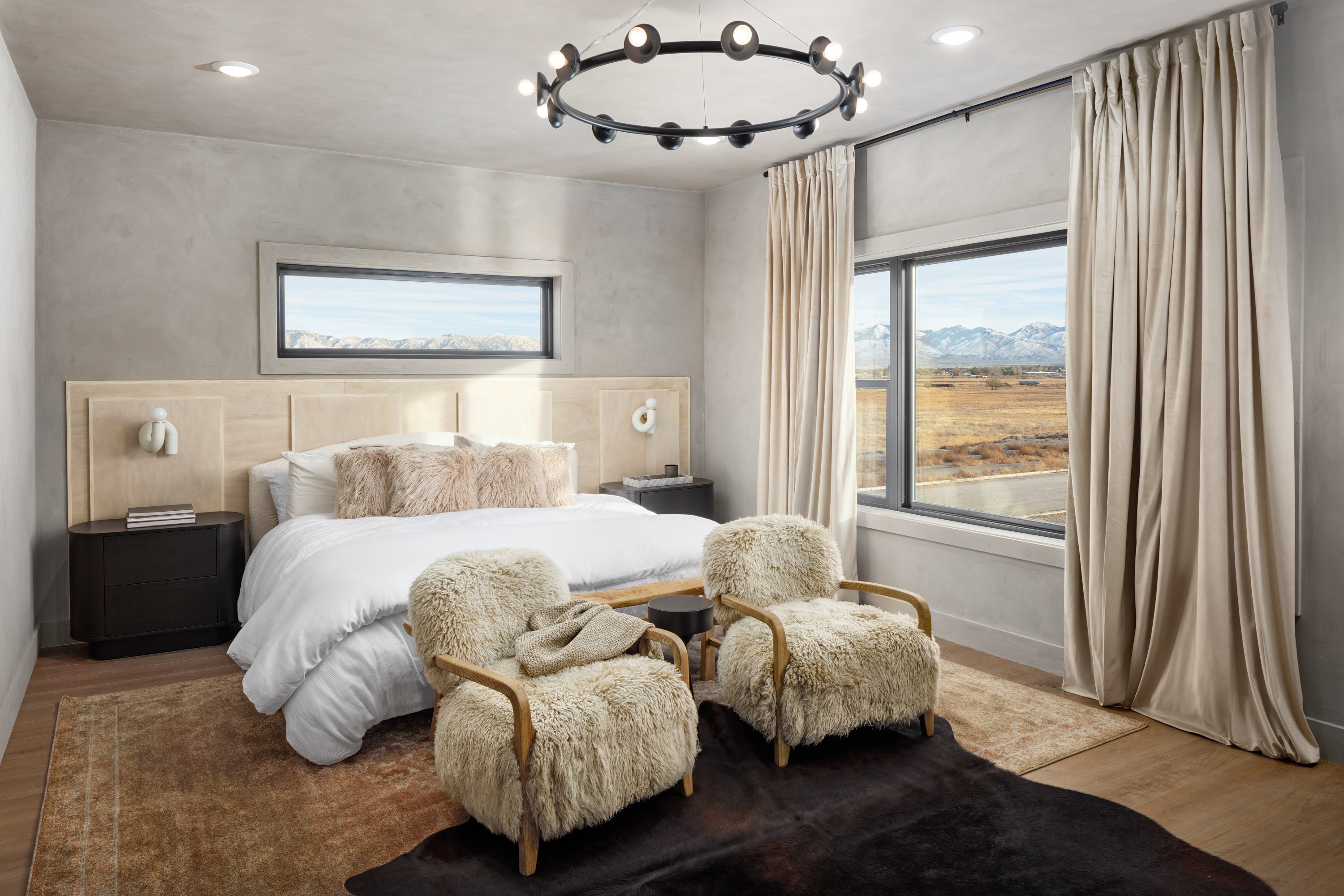 
      A primary bedroom with a bed made with a white duvet and neutral bedding accessories, as well as fluffy, fur lined chairs at the foot at the foot of the bed.
    