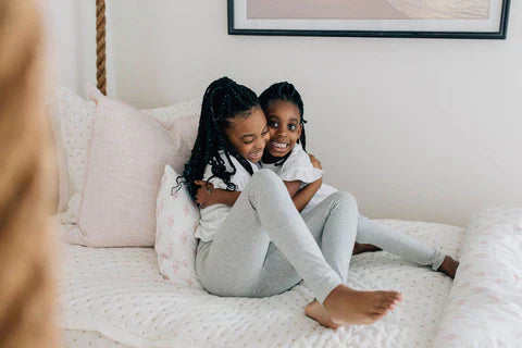 
      Two girls sitting and hugging on cream embroidered zipper bedding.
    