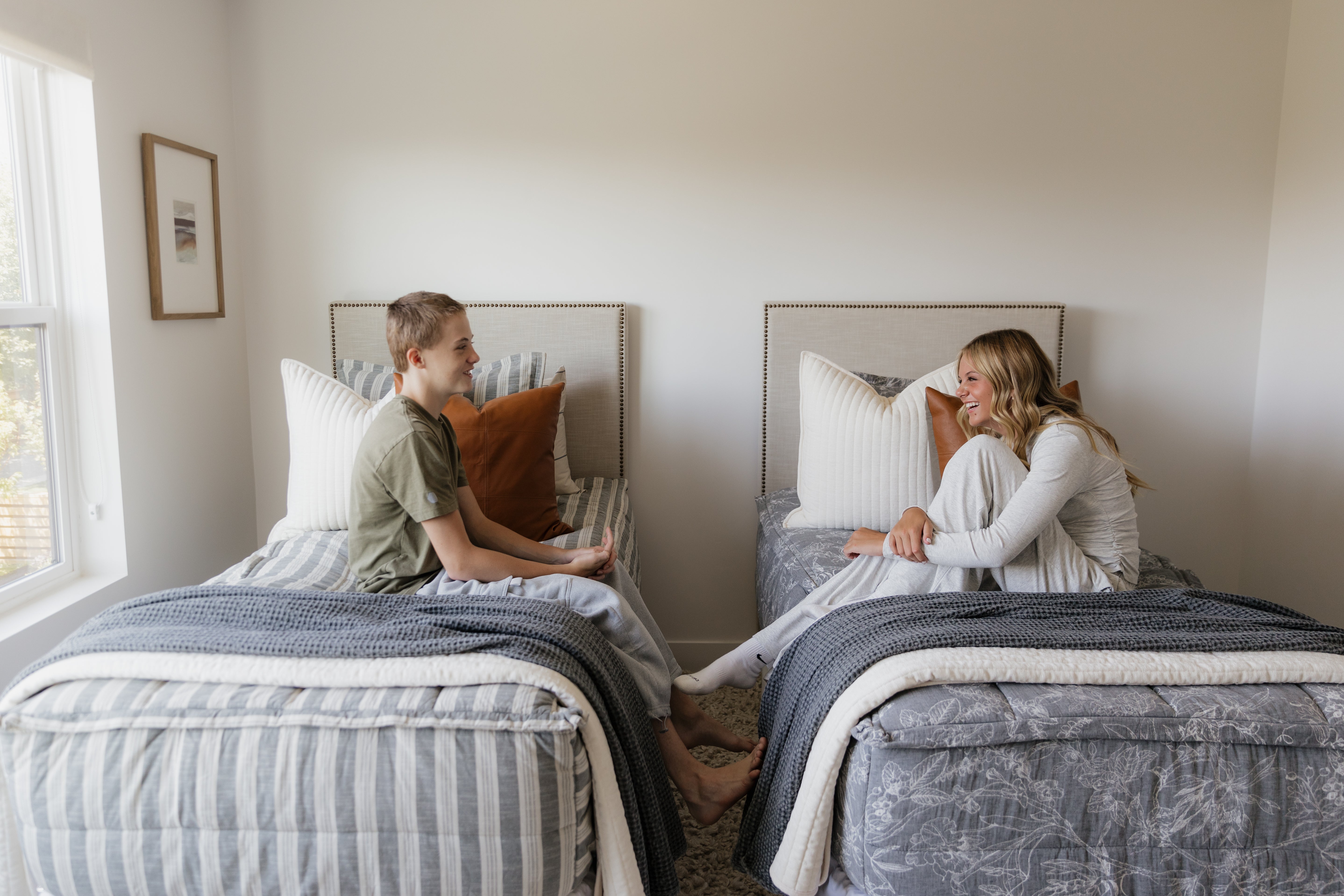 
      A bedroom with two twin beds, one made with striped zipper bedding and a teen boy sitting on top, the other made with floral zipper bedding and a teen girl sitting on top.
    