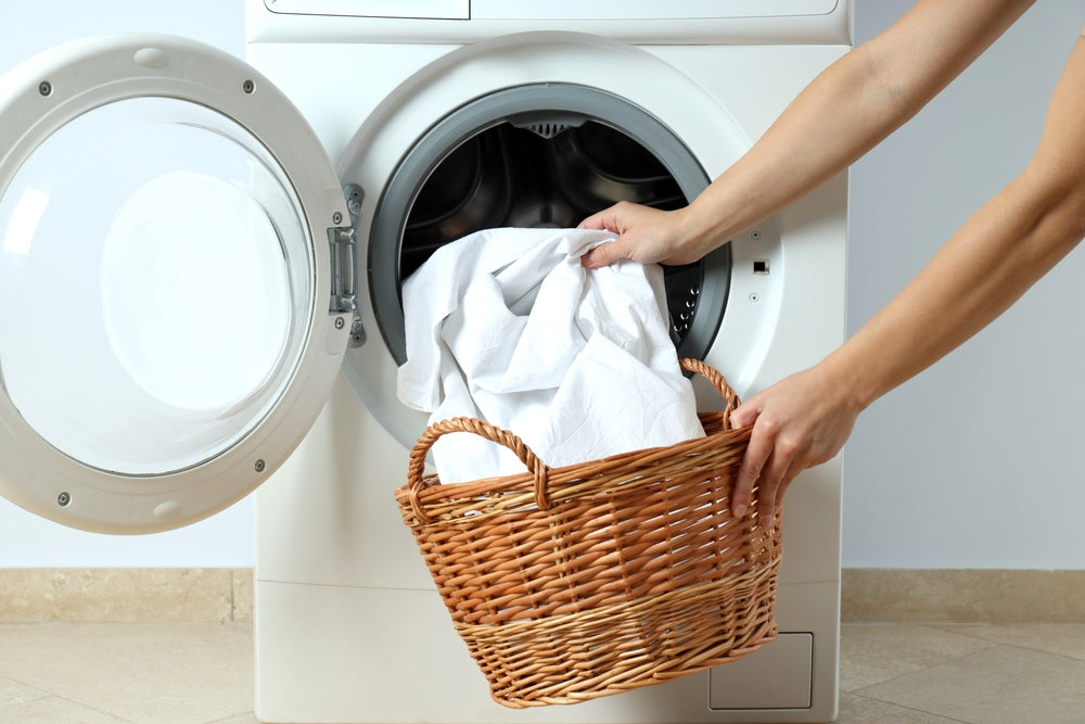 
      Someone opening a washer and pulling white bedding out and depositing it into a brown wicker basket.
    