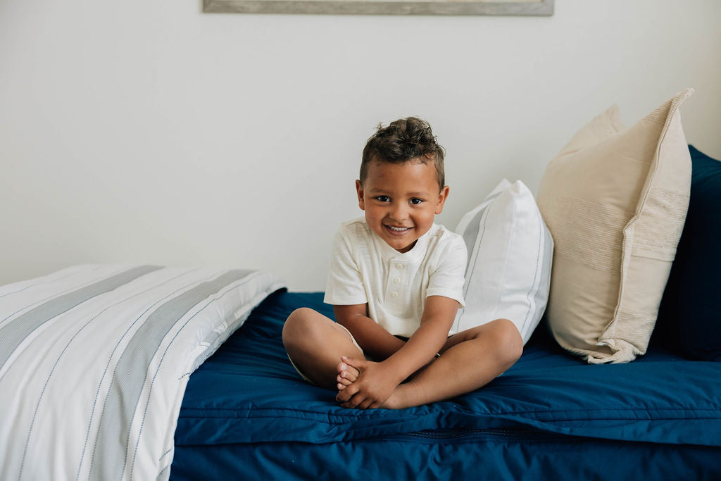 
      A young boy sitting cross legged on his bed, the bed made with navy blue zipper bedding and an assortment of neutral bedding accessories.
    