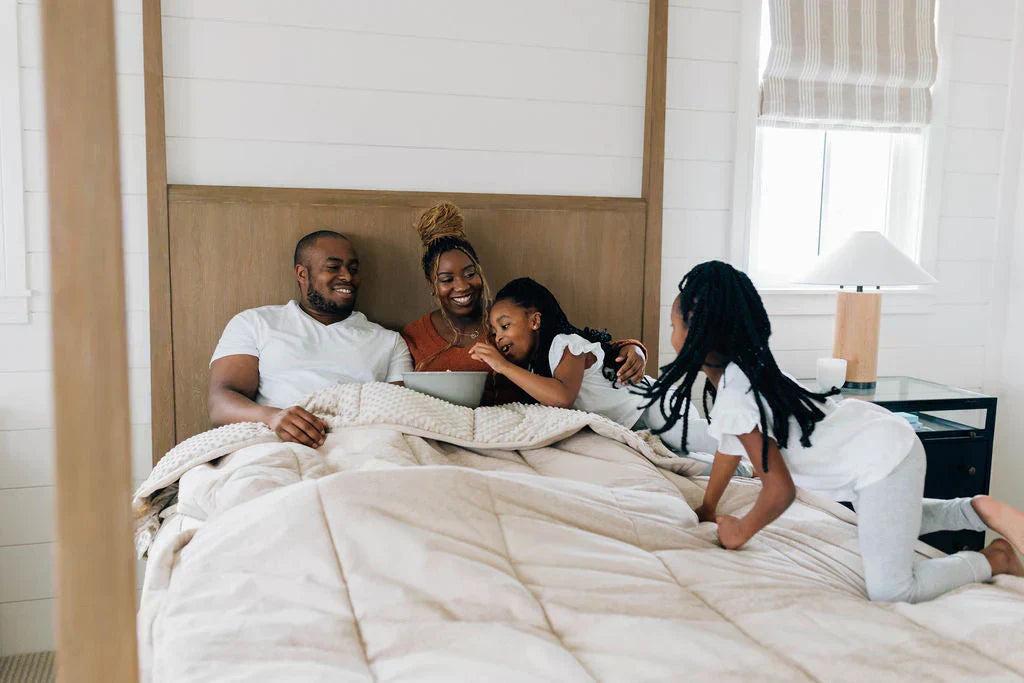 
      A husband, wife, and their two young daughters tucked in bed laughing together.
    